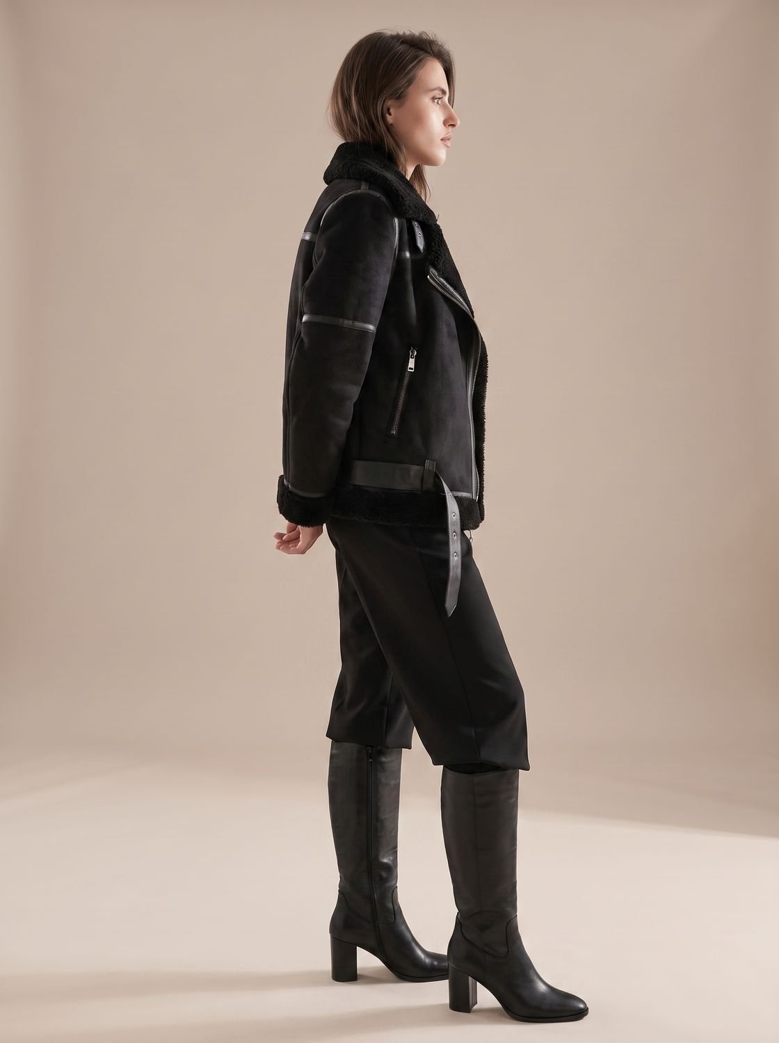 Black belted shearling aviator jacket for women worn by a model, featuring asymmetrical zip, plush lining, and wide collar