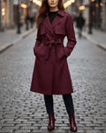 A woman wearing a burgundy belted trench coat with a double-breasted front and storm flap details, walking on a cobblestone street