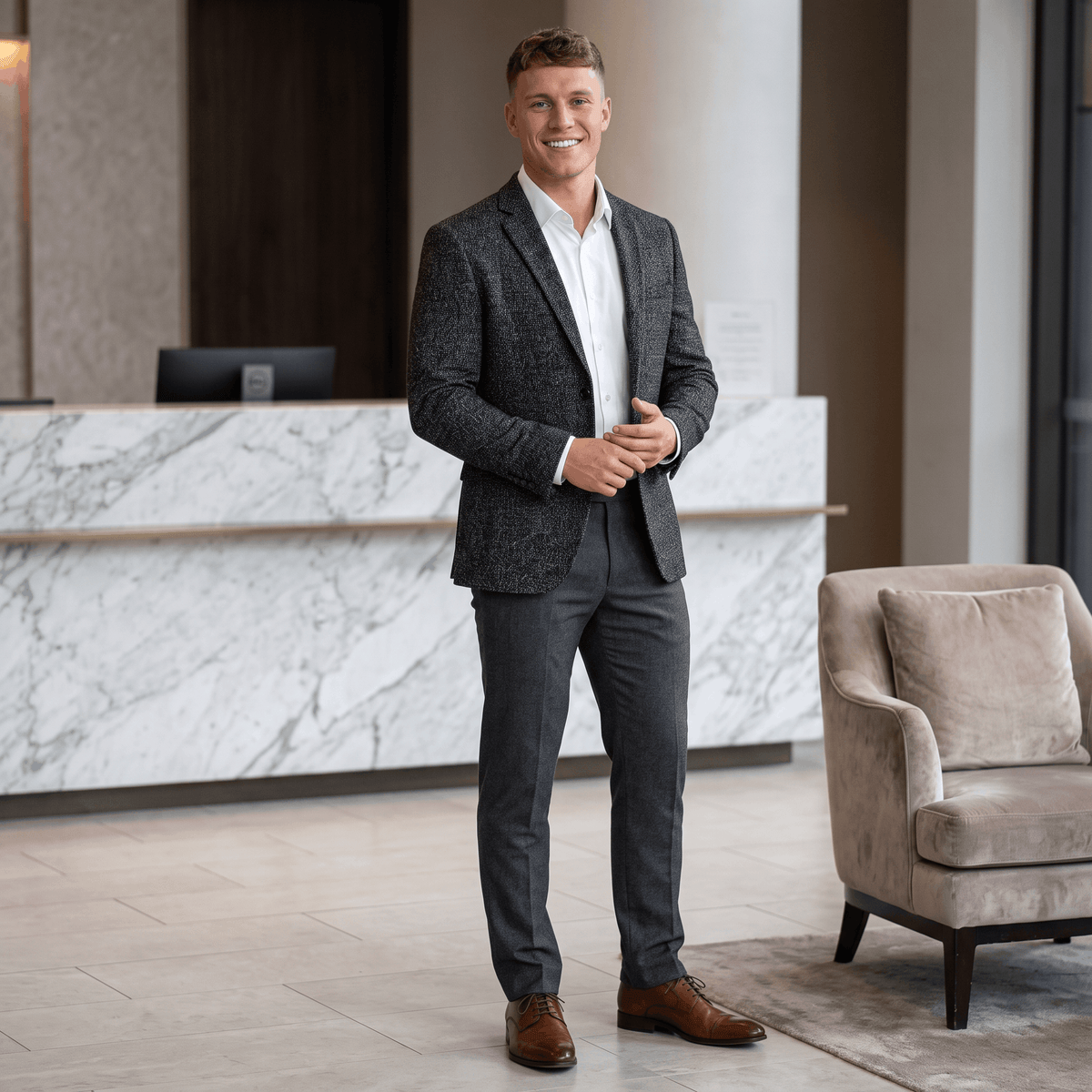 Man wearing a charcoal gray tweed blazer, white dress shirt, and dark trousers while standing in an elegant modern interior.