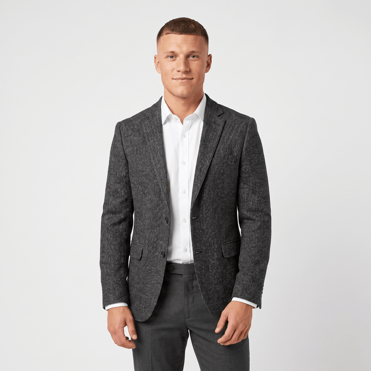 Man wearing a charcoal gray tweed blazer with a notched lapel, two-button closure, flap pockets, and tailored fit over a white dress shirt and dark trousers.