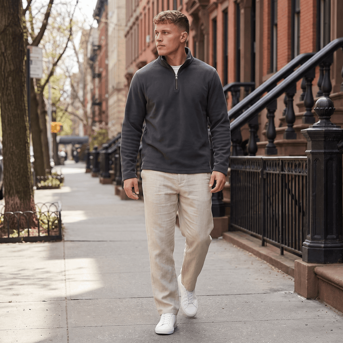 Man wearing a charcoal gray quarter-zip knit sweater with beige trousers walking along a city sidewalk.