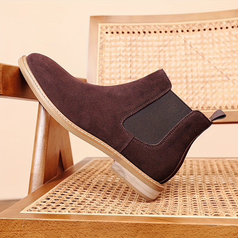 Dark brown suede Chelsea boot for men propped on a cane chair, showing the elastic side panel and pale sole