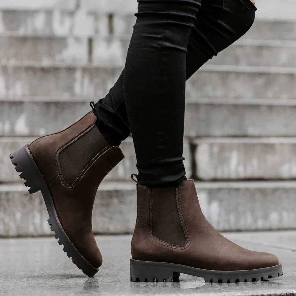 Dark brown chunky Chelsea boots for women on a lug sole, worn with black jeans on concrete steps