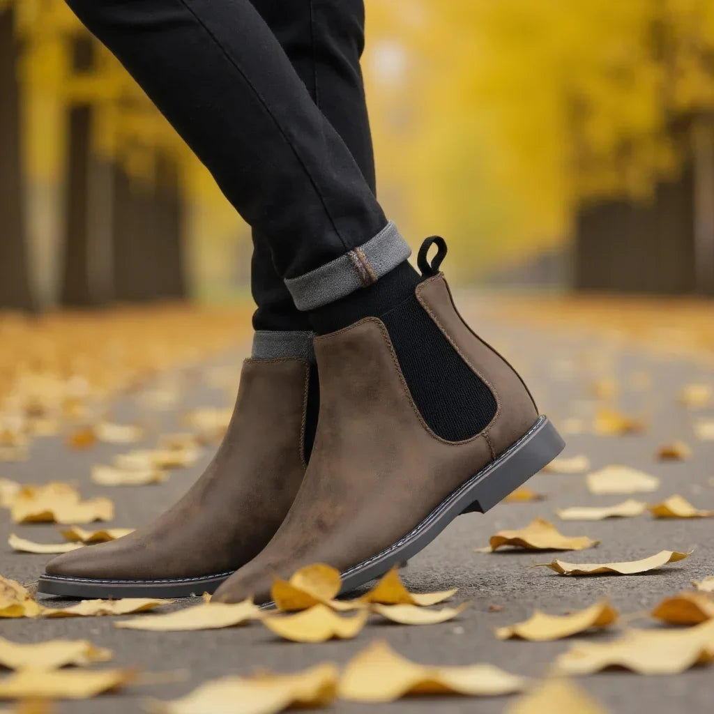 Distressed dark brown leather Chelsea boots with black elastic side panels and a stitched welt sole on an autumn path.