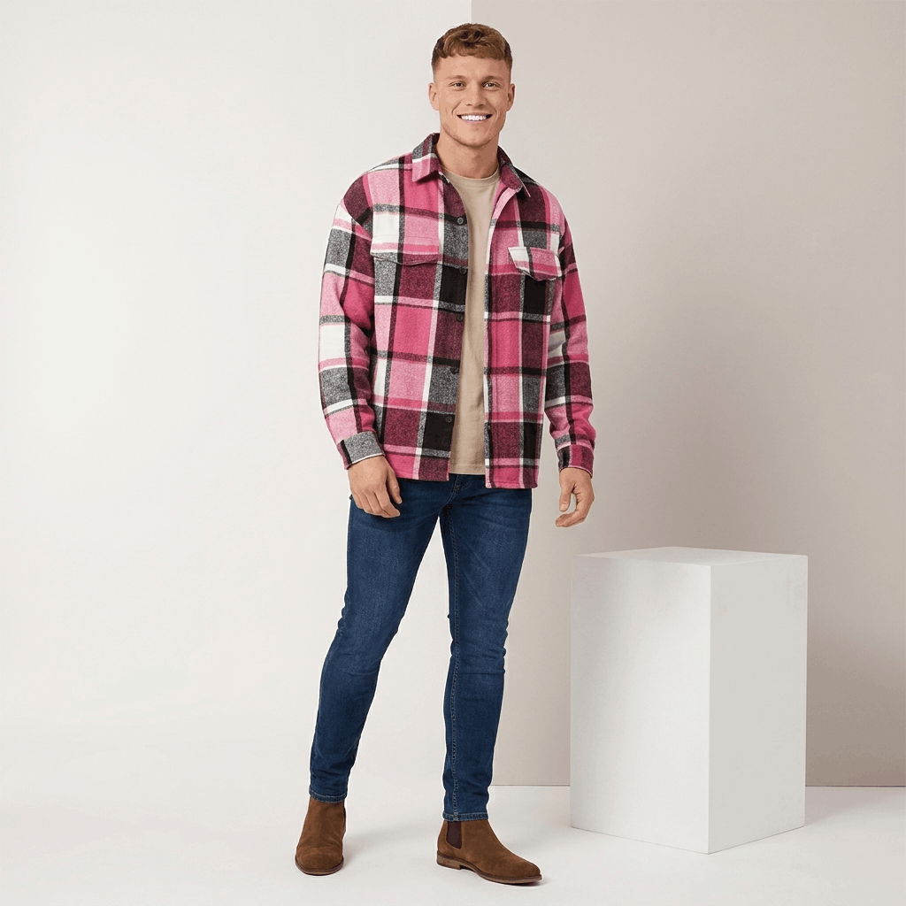 Man wearing a pink, burgundy, and white plaid overshirt jacket with beige trousers and brown shoes in a studio setting.