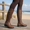 Smooth grain brown leather riding boots with a buckle calf strap and lug tread outsole worn over dark leggings.
