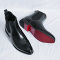 Pair of black leather Chelsea boots displayed with one boot showing its burgundy-colored sole, highlighting the sleek pointed design.