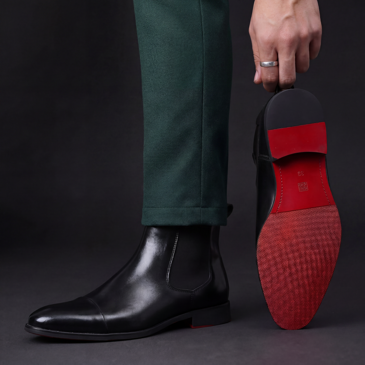 Man in green trousers wearing a black leather cap-toe Chelsea boot, holding the second boot up to show its red sole