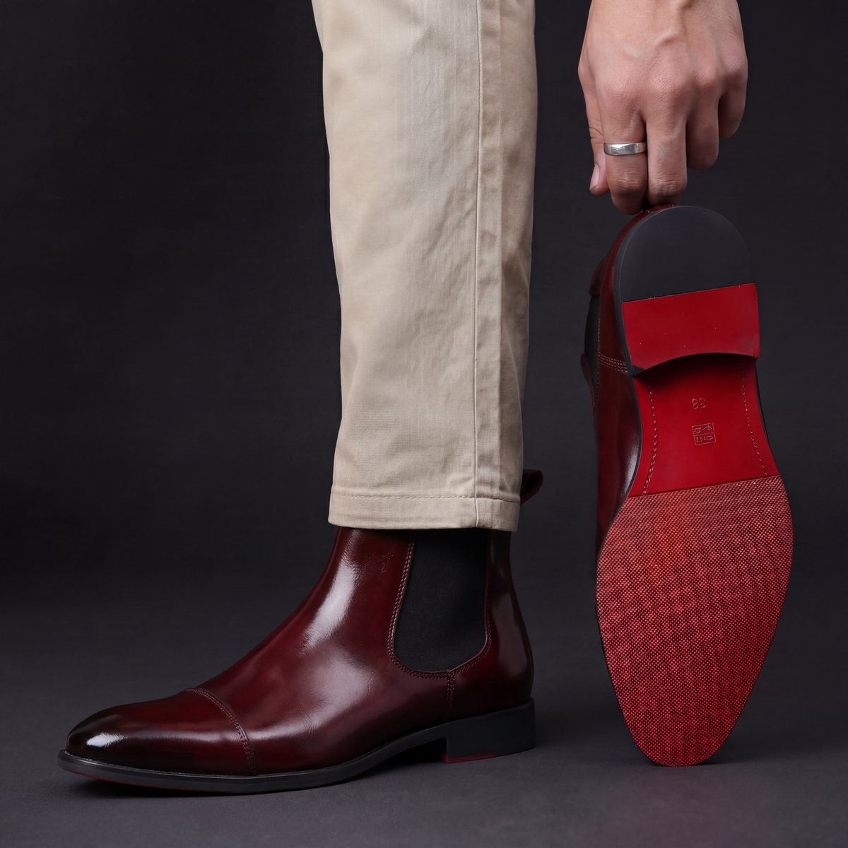 Man in beige chinos wearing a burgundy leather cap-toe Chelsea boot, second boot held up showing the red sole