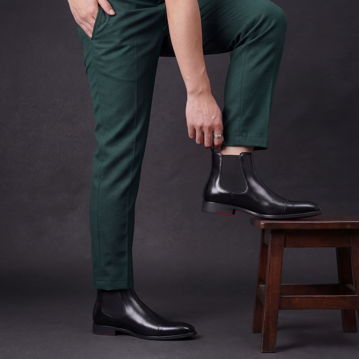 Man in green trousers resting one foot on a wooden stool, wearing black leather cap-toe Chelsea dress boots