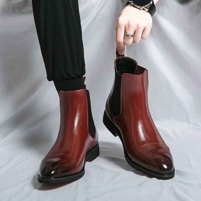 Man pulling on burgundy leather Chelsea boots by the heel tab, worn with black trousers on a white fabric backdrop