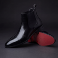 Black leather cap-toe Chelsea boots for men shown at an angle with red rubber sole visible, on a dark studio backdrop