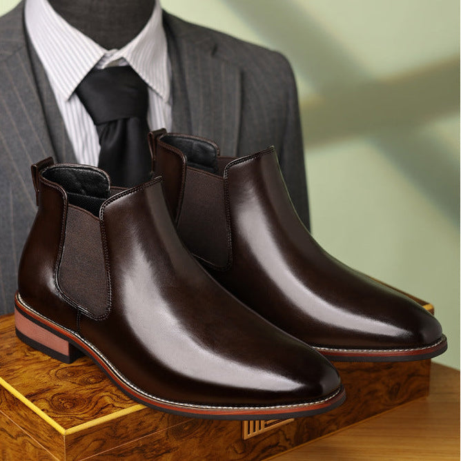 Pair of dark brown leather Chelsea boots displayed on a wooden surface with a suit in the background.