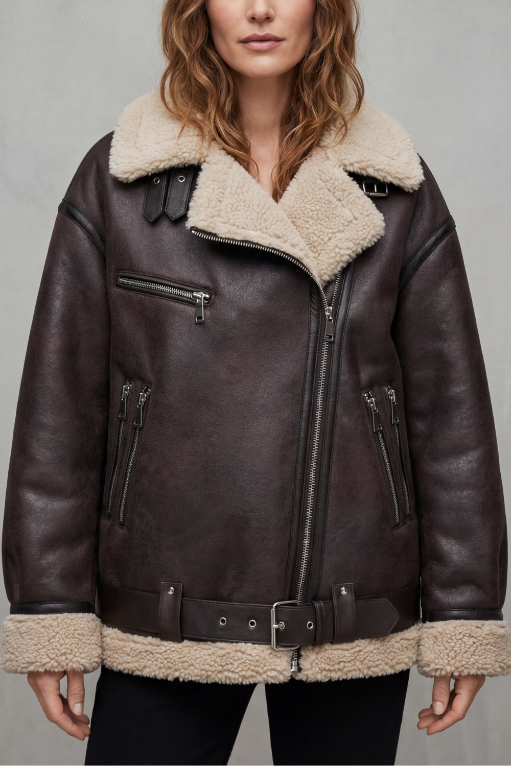 A woman wearing a dark brown leather shearling aviator jacket with a belted waist, zip front, and plush shearling collar and cuffs