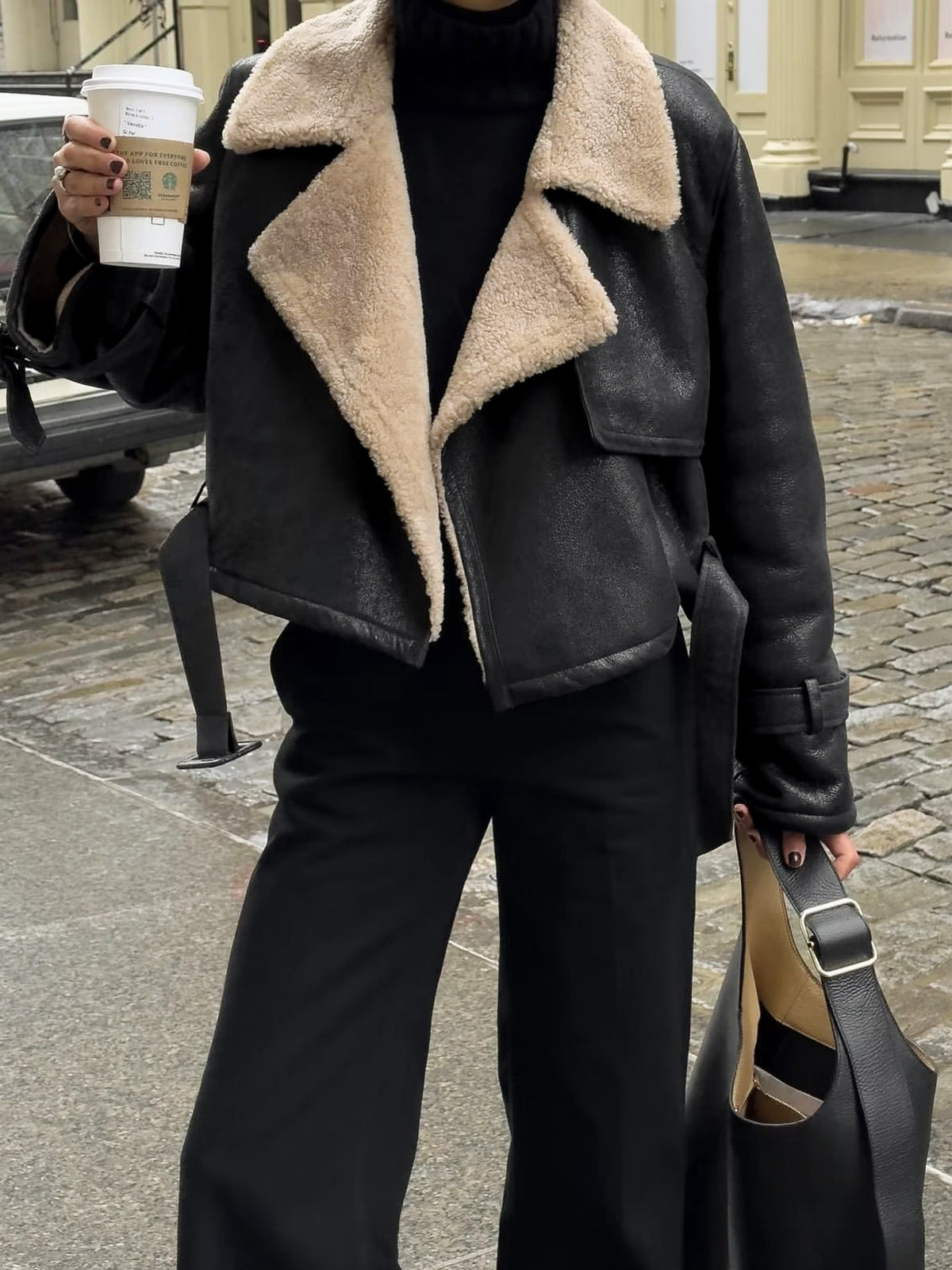 A woman wearing a black leather shearling aviator jacket with a cropped fit, plush shearling lining, and buckled cuffs, standing outdoors