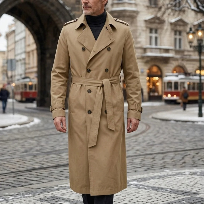 A man wearing a beige long double-breasted trench coat with an adjustable belt and tailored profile, standing on a city street