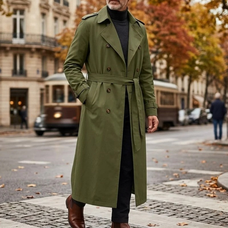 A man wearing an olive green long double-breasted trench coat with an adjustable belt and tailored profile, standing outdoors