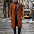 A man wearing a long brown double-breasted trench coat with an adjustable belt and tailored profile, standing on a city street