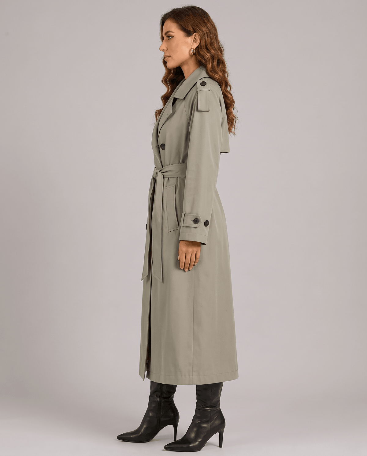 A woman wearing an olive green long trench coat with a double-breasted front and belted waist, shown in side view