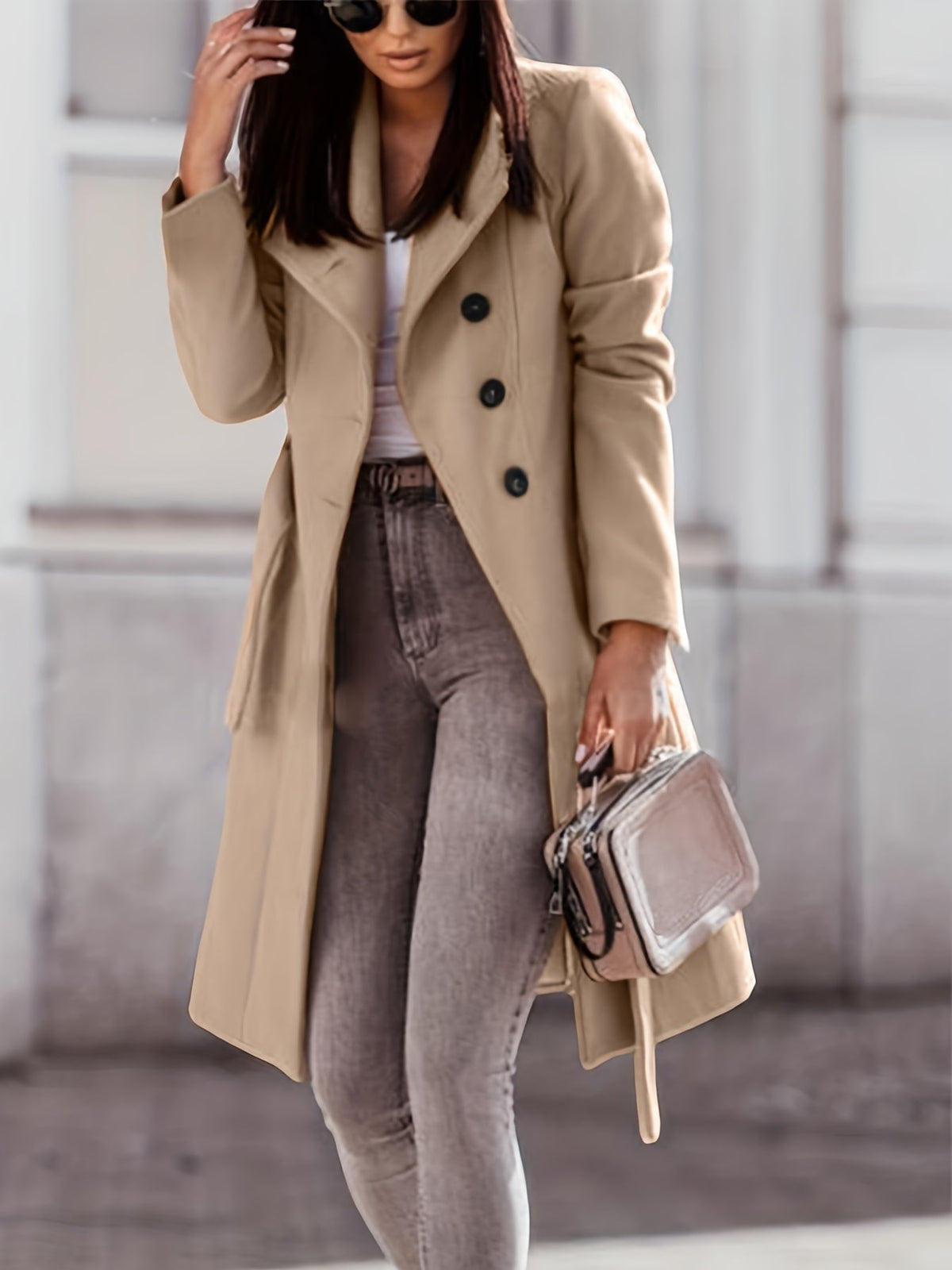 A woman wearing a beige long double-breasted trench coat with a belted waist, walking on a city street