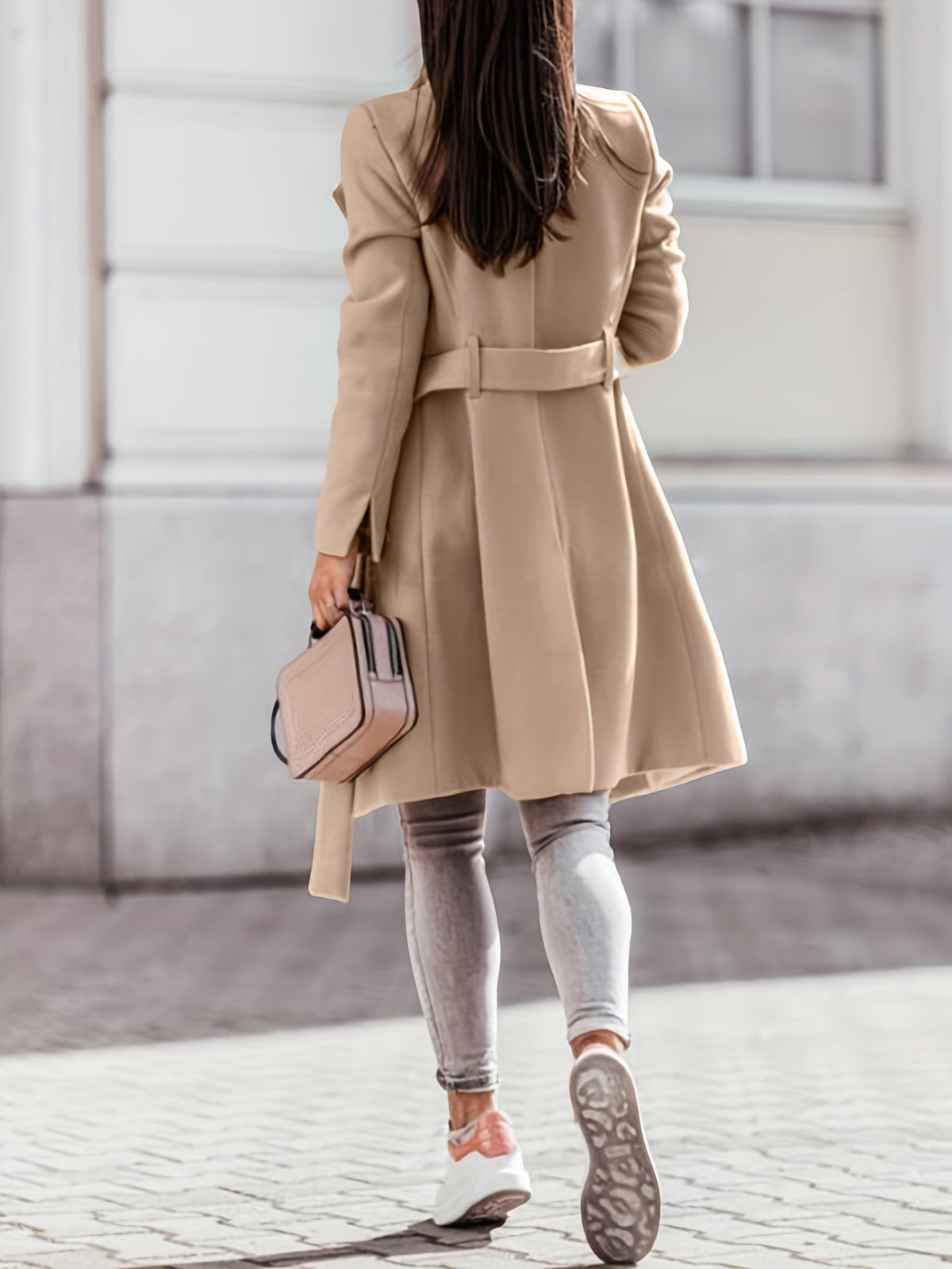 A woman wearing a beige long double-breasted trench coat with a belted waist, shown from the back