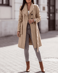 A woman wearing a beige long trench coat with a double-breasted front and belted waist, walking outdoors