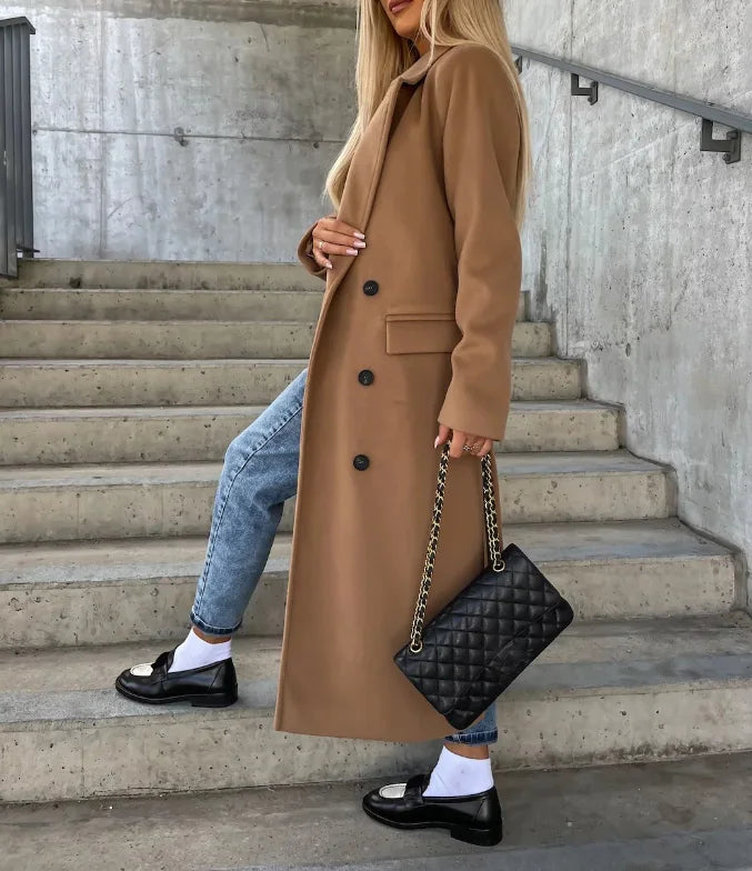 A woman wearing a long camel double-breasted trench coat with wide lapels and a belted waist, seated on stairs