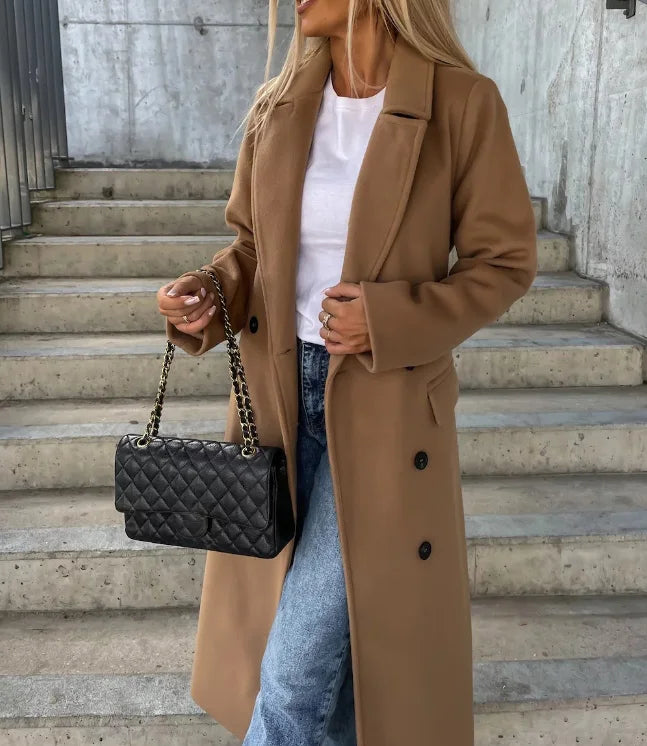A woman wearing a long camel double-breasted trench coat with wide lapels and a belted waist, holding a handbag