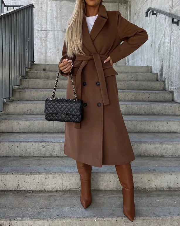 A woman wearing a brown double-breasted trench coat with wide lapels and a belted waist, carrying a handbag