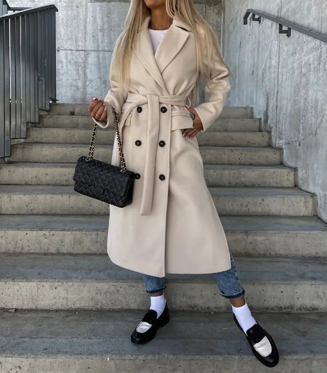 A woman wearing a white long double-breasted trench coat with wide lapels and a belted waist, standing on stairs