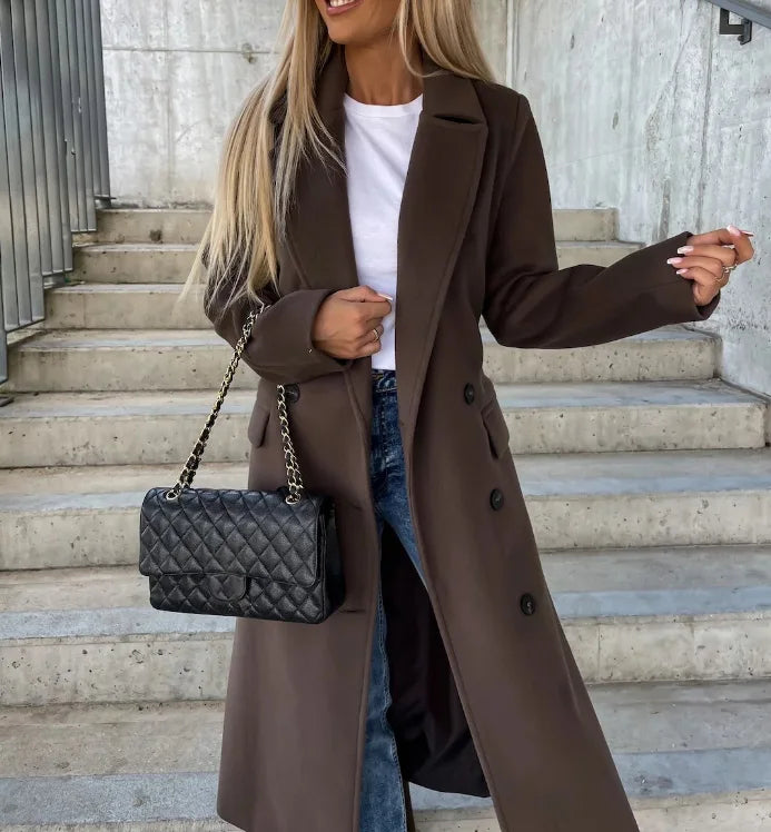 A woman wearing a dark brown long double-breasted trench coat with wide lapels and a belted waist, adjusting the coat