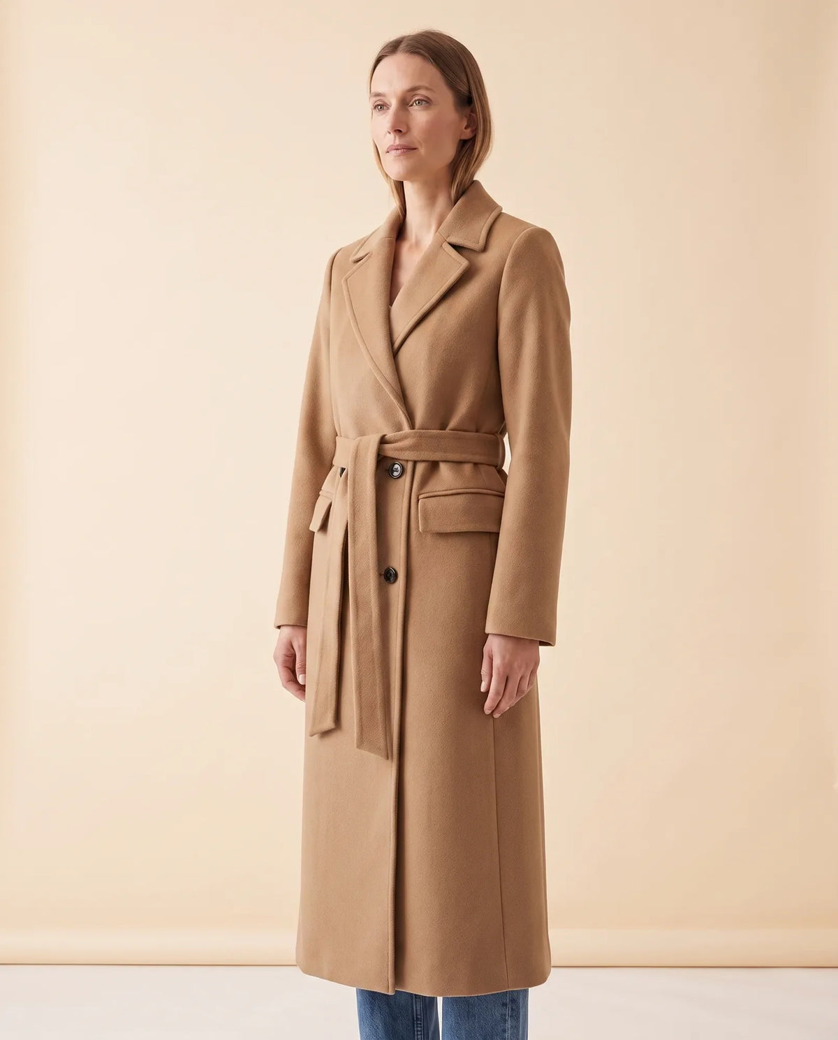 A woman wearing a camel longline belted coat with a double-breasted front and notched lapel, shown in side view