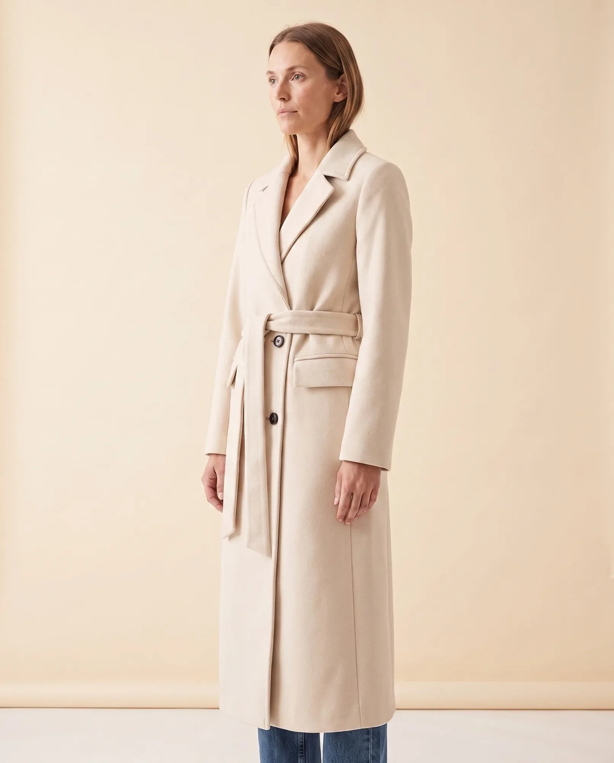 A woman wearing a cream longline belted coat with a double-breasted front and notched lapel, full-length view