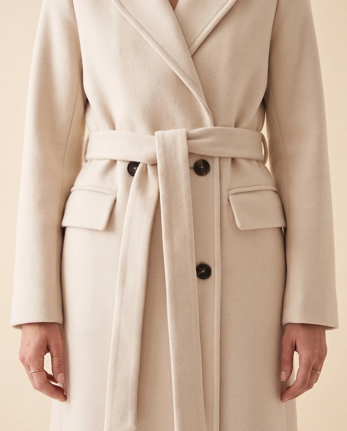 Close-up of a cream longline coat showing the double-breasted front, notched lapel, and belted waist