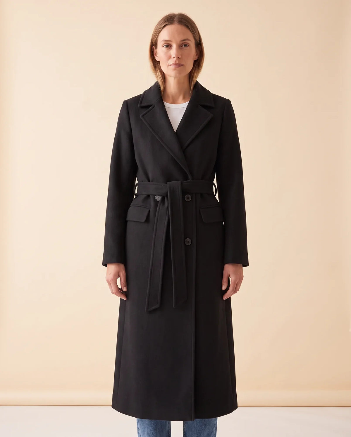 A woman wearing a black longline belted coat with a double-breasted front and notched lapel