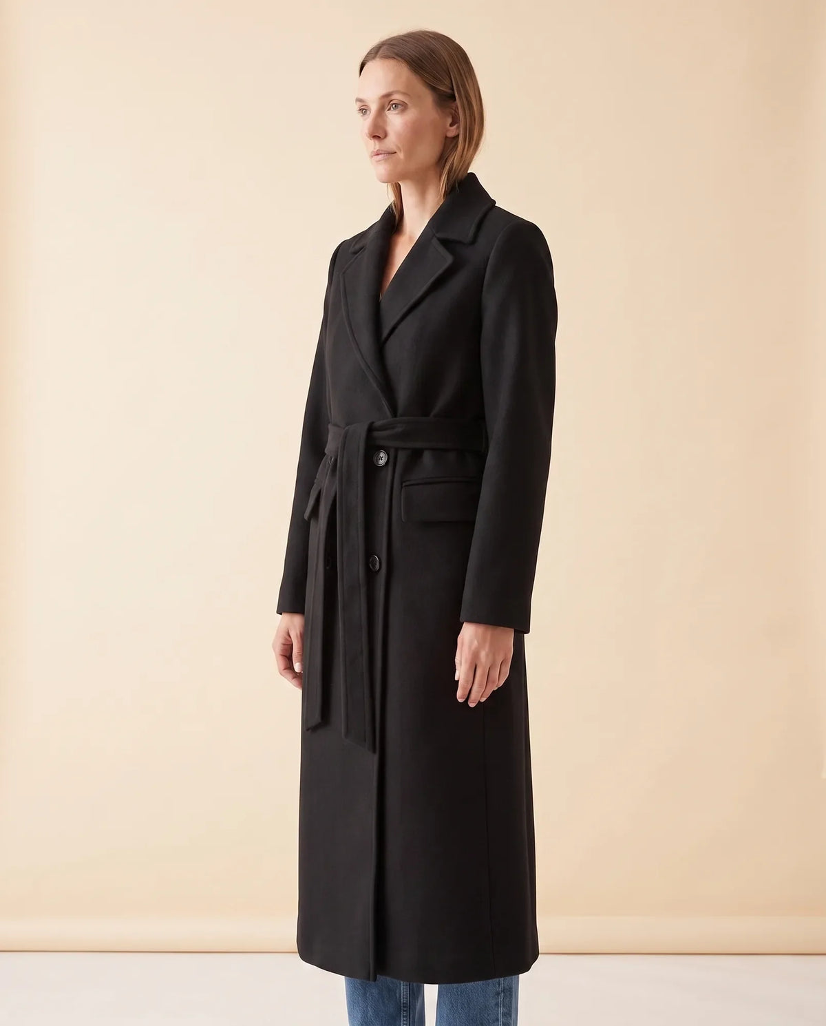 A woman wearing a black longline belted coat with a double-breasted front and notched lapel, shown in side view