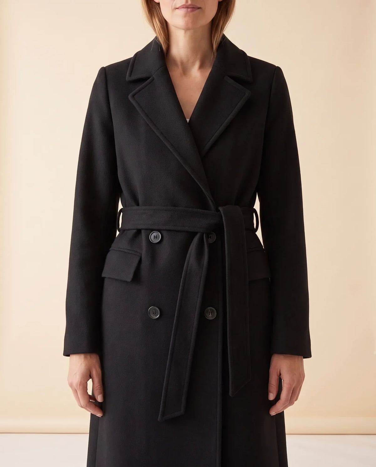 A woman wearing a black longline belted coat with a double-breasted front and notched lapel, front view
