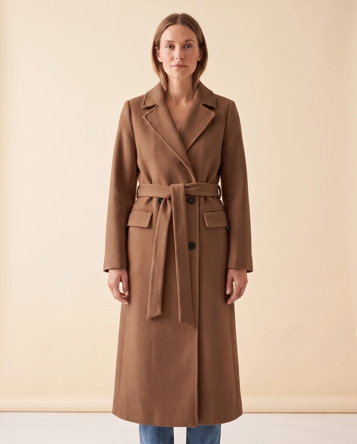 A woman wearing a brown longline belted coat with a double-breasted front and notched lapel