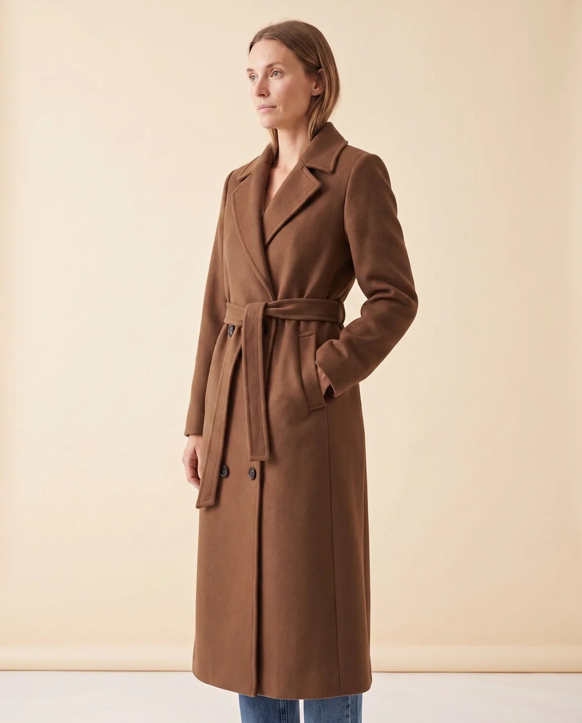 A woman wearing a brown longline belted coat with a double-breasted front and notched lapel, shown in side view