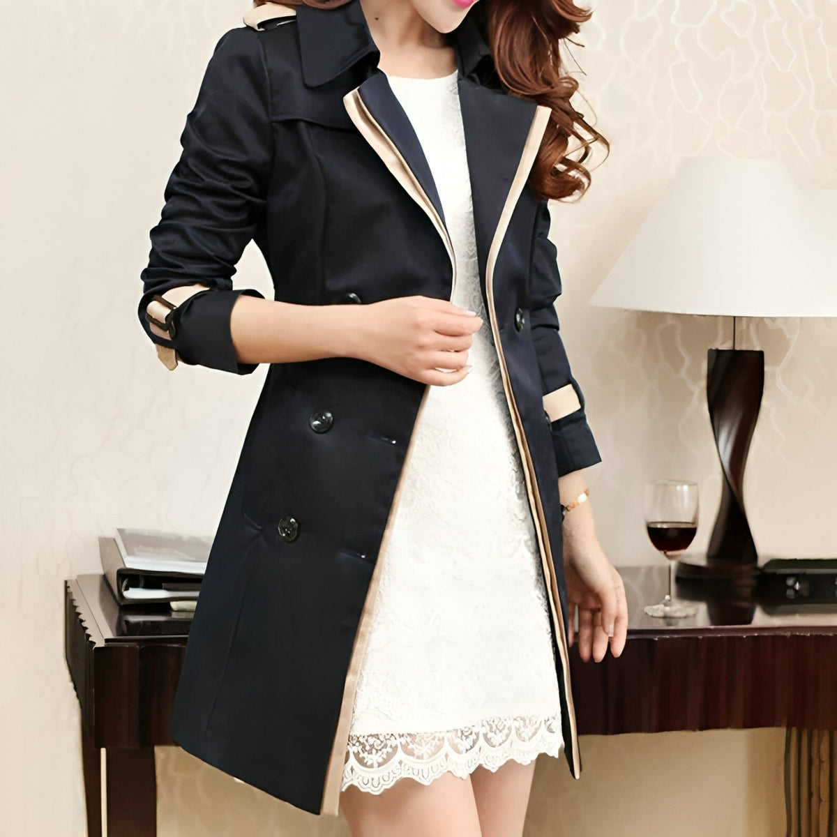 A woman wearing a black longline trench coat with a belted waist and double-breasted front, fastening the coat