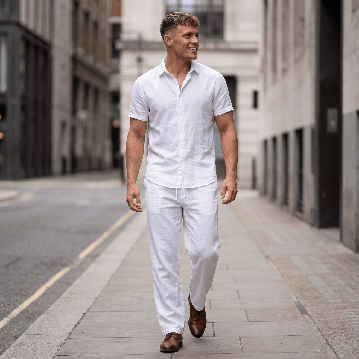 Man wearing white linen drawstring pants with a white linen shirt and brown leather sandals while walking through an urban street.