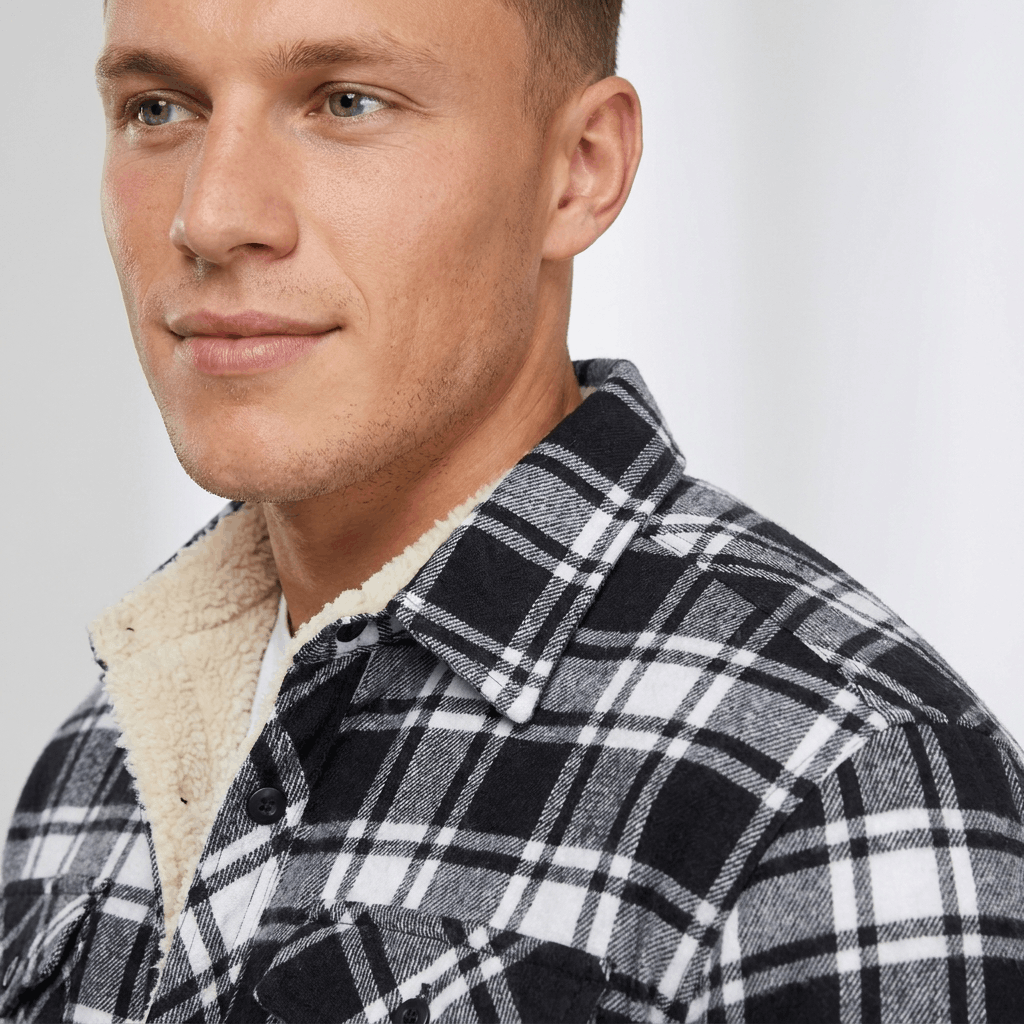 Close-up of a man wearing a black and white plaid sherpa-lined overshirt jacket, showing the pointed collar and textured plaid fabric.