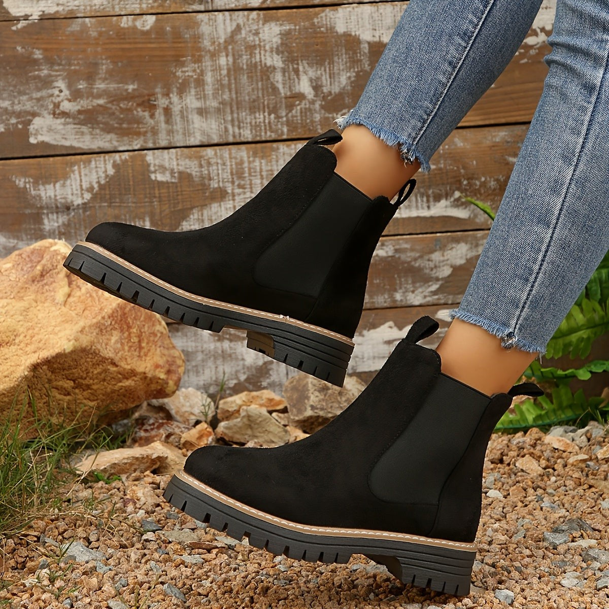 Black suede Chelsea boots for women, one foot lifted to show the lug tread and heel tab