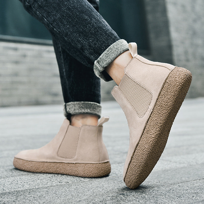Beige suede Chelsea sneaker boots with textured rubber soles and knit gore panels worn with dark cuffed jeans.