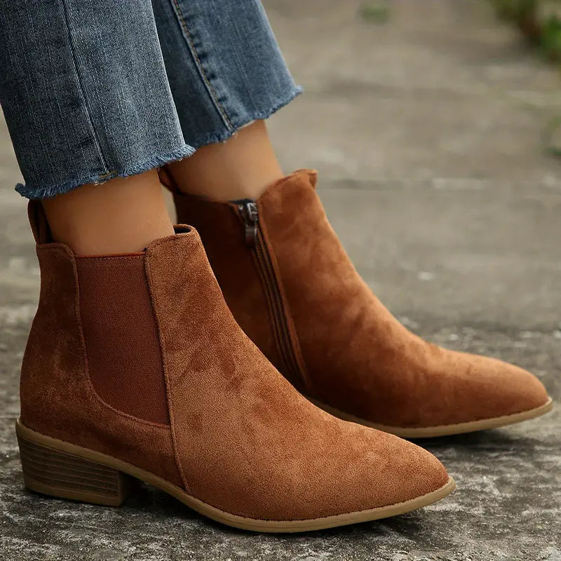 Brown suede Chelsea boots for women with pointed toe, one foot lifted to show the low stacked block heel