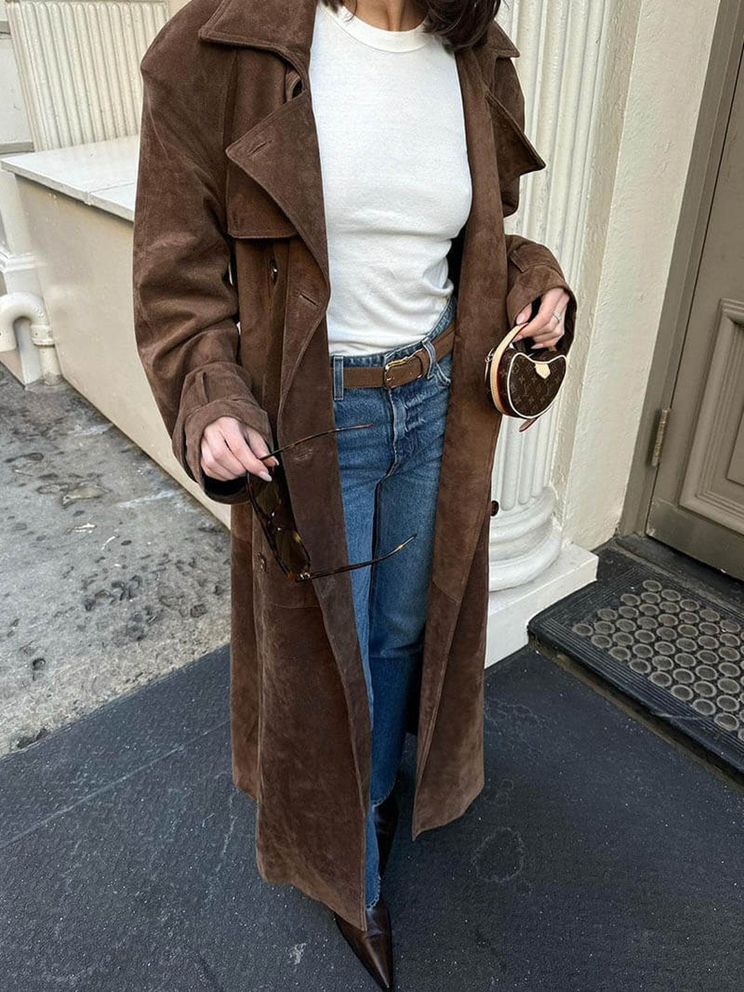 A woman wearing a brown suede trench coat with a belted waist, double-breasted front, and longline fit