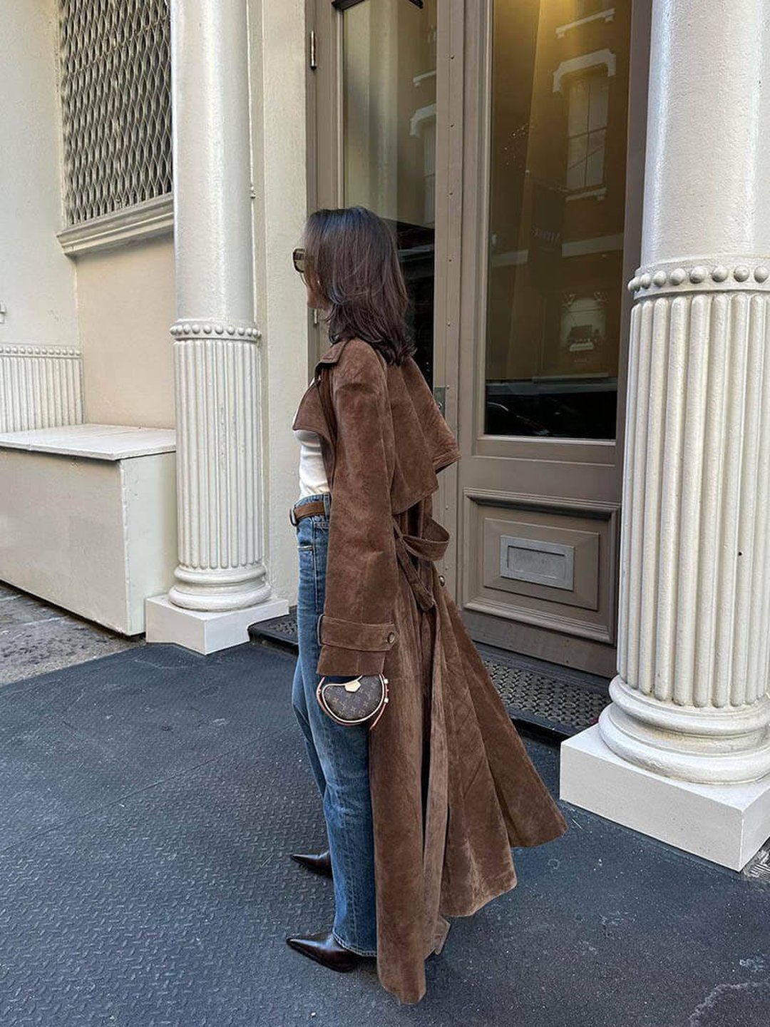 A woman wearing a brown suede trench coat with a belted waist and double-breasted front, standing outdoors