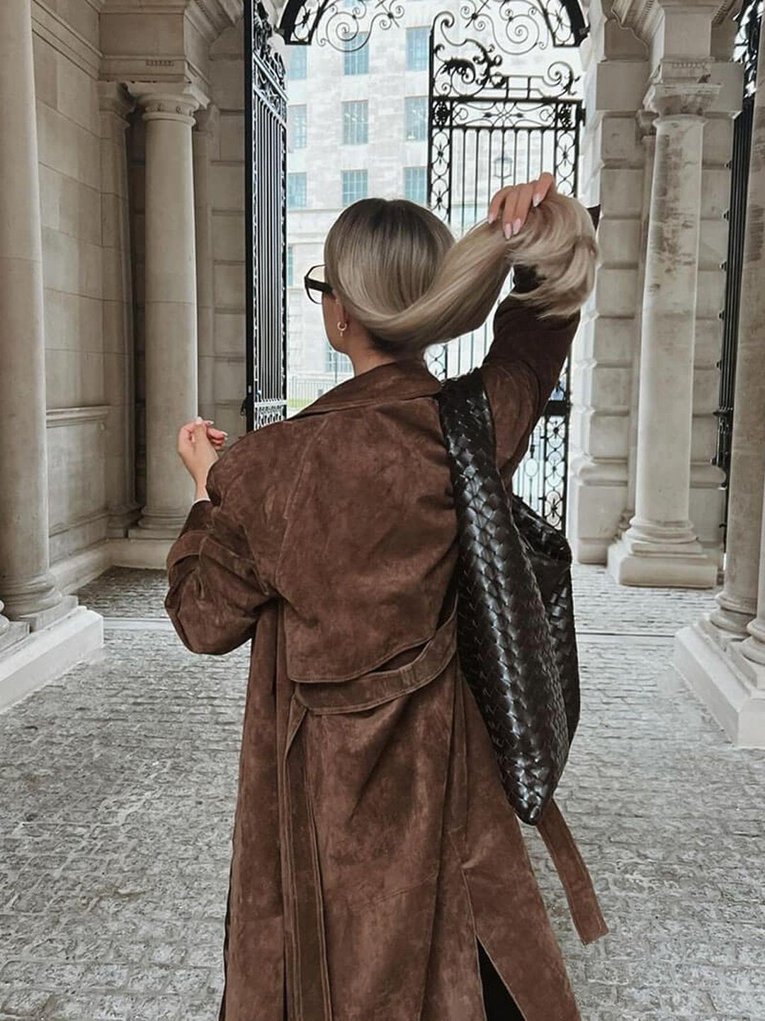 A woman wearing a brown suede trench coat with a belted waist and longline fit, viewed from the back outdoors