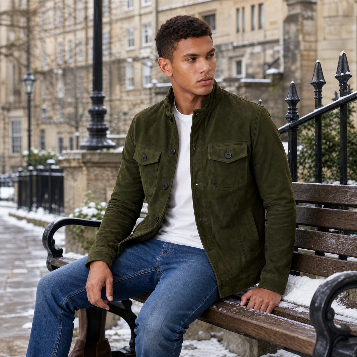 A man wearing an olive green suede trucker jacket with a button front, stand collar, and chest pockets, seated outdoors