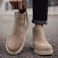 Man pulling on a beige vegan suede Chelsea boot by the rear tab, worn with cuffed black jeans